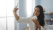 © Krakenimages.com - Young woman taking a selfie holding a beauty product in a spa room, wearing a robe, surrounded by wellness items, showcasing a serene indoor setting.