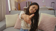 © Krakenimages.com - Beautiful latina woman with long brunette hair smiling and pointing at the camera while sitting on a comfortable sofa in a cozy living room apartment