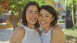 © Krakenimages.com - Two smiling women who appear to be hispanic stand closely together outdoors in a city park, with trees and urban elements visible in the background.