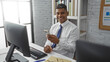 © Krakenimages.com - Young hispanic man smiling in office looking at smartphone seated at desk with computer and bookshelves in background