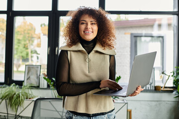 Wall Mural - A young woman stands confidently with a laptop in a bright, plant filled workspace.