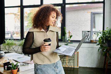 Wall Mural - A woman engages with her laptop, holding a coffee cup in a contemporary office filled with greenery.