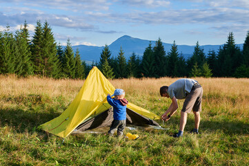 Wall Mural - Man hiker and young boy work together to set up tent on grassy hillside. Pine trees and distant mountains under partly cloudy sky.