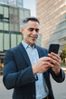 © Jose Calsina - Vertical. Mature businessman using cellphone outdoors, professional man with gray hair and beard in suit looking at phone, standing confidently in urban area engaging with technology and communication