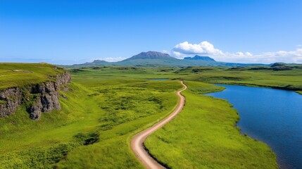 Wall Mural - winding dirt path meanders through lush green fields next to a serene lake, with majestic mountains rising dramatically against a backdrop of clear blue skies