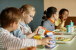 © Seventyfour - Side view of young girl pouring fresh juice or milk into cup sitting at table with classmates while eating lunch in school cafeteria, copy space