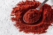 © Kanjana - A spoonful of vibrant red spice rests on a scattered pile, showcasing its rich texture and color against a light background.