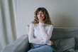 © wifesun - Young woman with curly hair wearing casual clothes is covering her mouth with hand while sitting on comfortable sofa in living room, expressing surprise or shock
