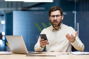 Portrait of an upset and disappointed young man in a shirt and glasses sitting in the office at the desk, holding the phone and spreading his hands to the camera