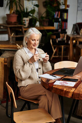 A beautiful mature woman with gray hair savors a drink, immersed in her work at a cafe.