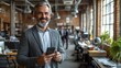 © Bagas - A smiling man in a modern office setting holds a smartphone while interacting with others.