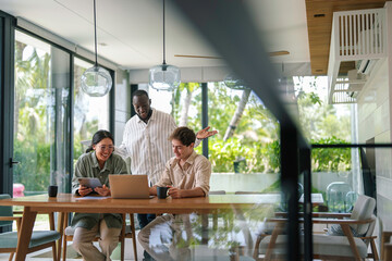 Wall Mural - Three young adult business professionals, of varied ethnicities, collaborate using a laptop and digital tablet in a sunlit modern office space.