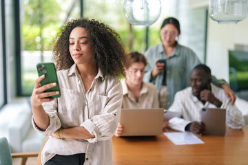 Wall Mural - A cheerful African American businesswoman engages with her smartphone, surrounded by diverse colleagues working on laptops in a well-lit modern office setting.