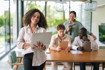 Wall Mural - A joyful African American businesswoman works on her laptop, standing confidently in a modern office, with diverse colleagues engaged in tasks behind her.