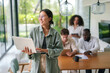 © Dorde - A cheerful Asian young adult businesswoman engages with her laptop, dressed in casual work attire, in a well-lit modern office as diverse colleagues collaborate in the background.