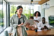 © Dorde - An Asian young adult female in casual business attire enjoys her work on a laptop, standing in a well-lit modern office with diverse colleagues in the background.