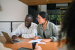 © Dorde - A young adult Black male and Asian female executive work together at a laptop in an office setting, discussing strategies with focused attention.