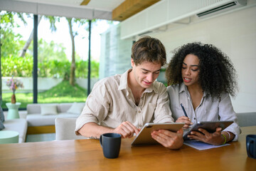 Wall Mural - African American woman and Caucasian man in casual attire engage with a digital tablet while sitting at a wooden table, with a bright home interior as the backdrop.