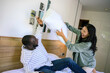 © Dorde - A cheerful Black man and Asian woman, both young adults, playfully engage in a pillow fight, expressing joy in casual attire in their cozy bedroom.