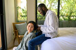 © Dorde - A cheerful Asian woman is seated as her smiling Black partner stands behind her, both dressed in casual attire, sharing a peaceful moment together in a sunlit cozy bedroom.