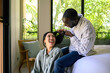 © Dorde - A Black man styles a cheerful Asian woman's hair, both dressed in casual attire, sharing an intimate moment in a sunlit room, surrounded by greenery.