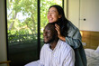 © Dorde - A cheerful Asian woman embraces a smiling Black man from behind, displaying a moment of domestic joy in a sunlit room with nature visible outside.