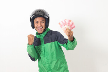 Portrait of Asian online taxi driver wearing green standing against white background, holding a bunch of rupiah money while raising fist, celebrating success