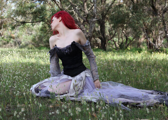 full length portrait of beautiful red haired female model figure, wearing gothic fantasy costume with torn ballgown wedding dress, black corset. sitting pose in flower field forest scenery background