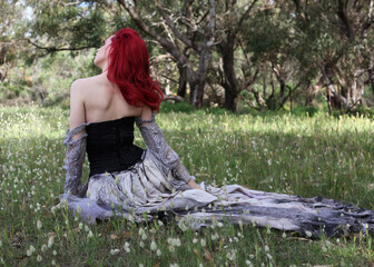 Wall Mural - full length portrait of beautiful red haired female model figure, wearing gothic fantasy costume with torn ballgown wedding dress, black corset. sitting pose in flower field forest scenery background