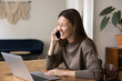 © fizkes - Pretty woman sit at desk with laptop, laughing, lead pleasant conversation on smartphone, discuss task with colleague, speaking with customer services support while making on-line order. Communication
