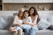 © fizkes - Portrait of pretty three generational family women posing for camera resting together on cozy couch. Lovely little girl enjoying priceless time with older granny and young mommy at home. Ties, love