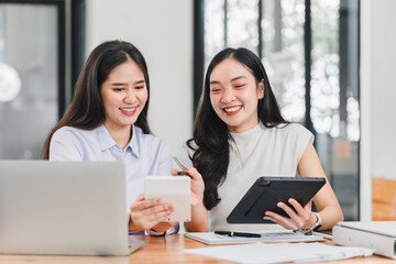 Wall Mural - Two women collaborating at desk, using smartphone and tablet, smiling and engaged in discussion. modern workspace features laptop and documents, creating productive atmosphere