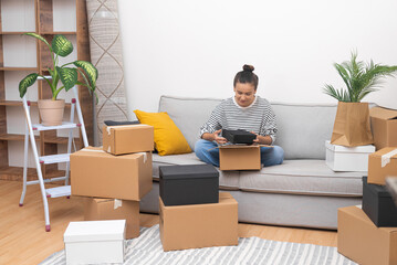 Aufkleber - Smilling lady with bun o head with delighted expression sits on sofa collecting cardboard boxes for moving in new apartment with all modern furniture
