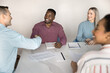 © fizkes - Diverse male business partners shaking hands at large office table shared with team, meeting for negotiation, closing deal, discussing partnership, project investment, giving handshake, smiling