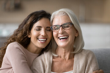 Wall Mural - close up of elderly mother and millennial grown-up daughter, laughing, cuddling seated together on c