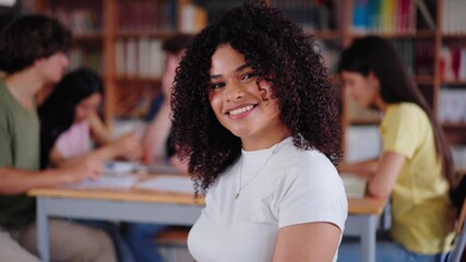 Wall Mural - High school girl poses looking smiling at camera sitting in library with classmates unfocused in background. Group friends meeting together in study room. Portrait happy young Latin female student