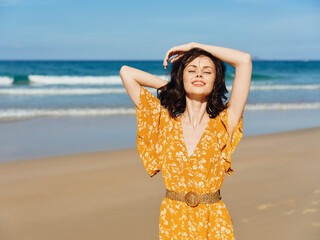 Poster - Woman in Yellow Dress Standing on Beach with Hands on Head, Enjoying Serene Ocean View