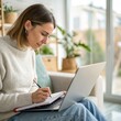 © SerPak - A young woman writes in a notebook while using a laptop in her bright living space.