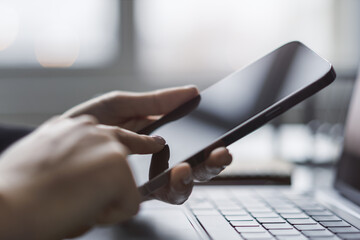 Wall Mural - Woman expertly using a cutting-edge smartphone at her workspace, laptop on standby against the vague backdrop of a thriving office