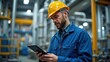 © useful pictures - A man wearing a hard hat and blue work jacket stands in a factory, focused on his tablet. The environment features large machinery and pipes, indicating an industrial setting