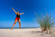 © Jacek Chabraszewski - Young woman in one piece black swimsuit, orange pareo and sunglasses standing with her legs apart and her arms raised up on white sand on beautiful beach on sunny summer day. Energy. Front view