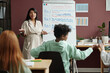 © pressmaster - Young confident female teacher looking at schoolchildren while standing in front of whiteboard with grammar rules