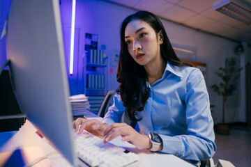 Wall Mural - Young asian businesswoman concentrating while using desktop computer, working overtime at night in modern office with blue light