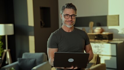 Canvas Print - Happy mid adult man in his 50s working on laptop computer standing in modern living room. Portrait of businessman managing business online in home office, smiling.