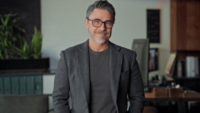 Fototapete -  - Confident mature businessman standing in office. Entrepreneur managing business online Portrait of happy mid adult, middle aged man in glasess with gray hair, smiling.