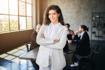 Fototapete - A woman in a light suit confidently poses in a contemporary office, with her arms crossed. Colleagues engage in discussion at a table, highlighting teamwork and collaboration.