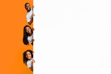 Discount. Smiling young black family of three holding white advertisement board, presenting free copy space for your text or design, positive people peeking out blank sign board banner, yellow studio