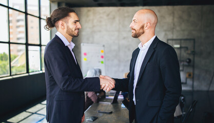 Aufkleber - Two men in suits greet each other with a handshake in a contemporary office. A group of colleagues is engaged in discussion in the background, emphasizing collaboration and professionalism.