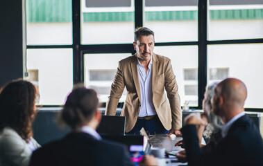 Fototapete - A business professional stands at a conference table, addressing a team during an important meeting in a contemporary office setting. Members actively participate and listen intently.