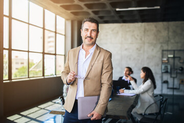 Aufkleber - A confident business professional stands in a modern office, holding documents and glasses. Colleagues engage in discussion at a table, showcasing teamwork and collaboration.
